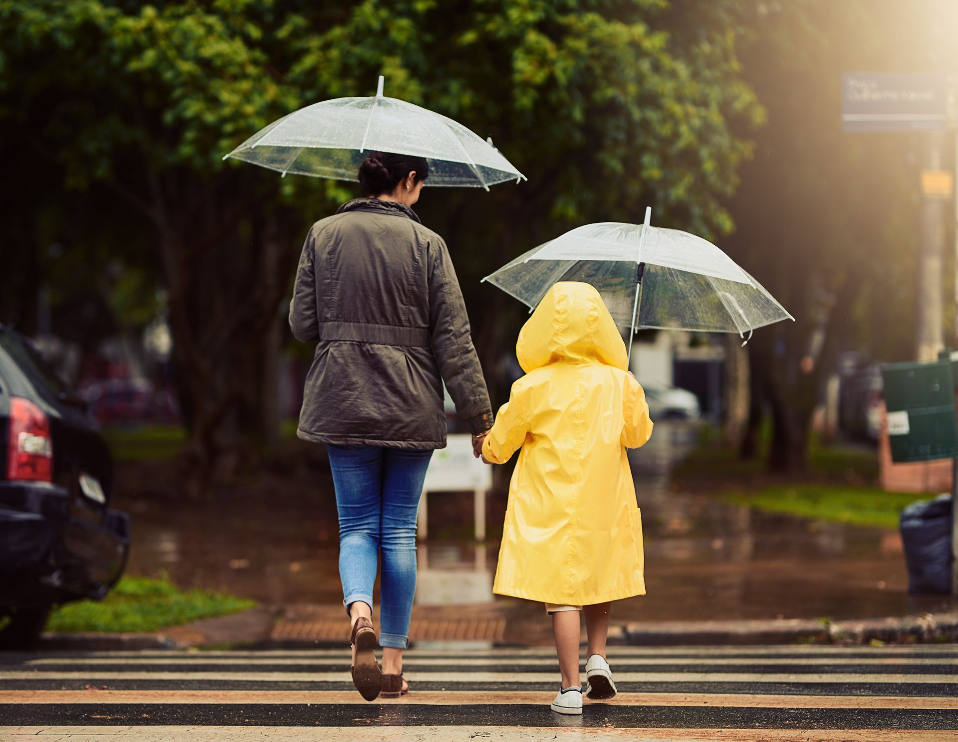 Woman and child in rain gear holding hands, crossing a street with umbrellas.