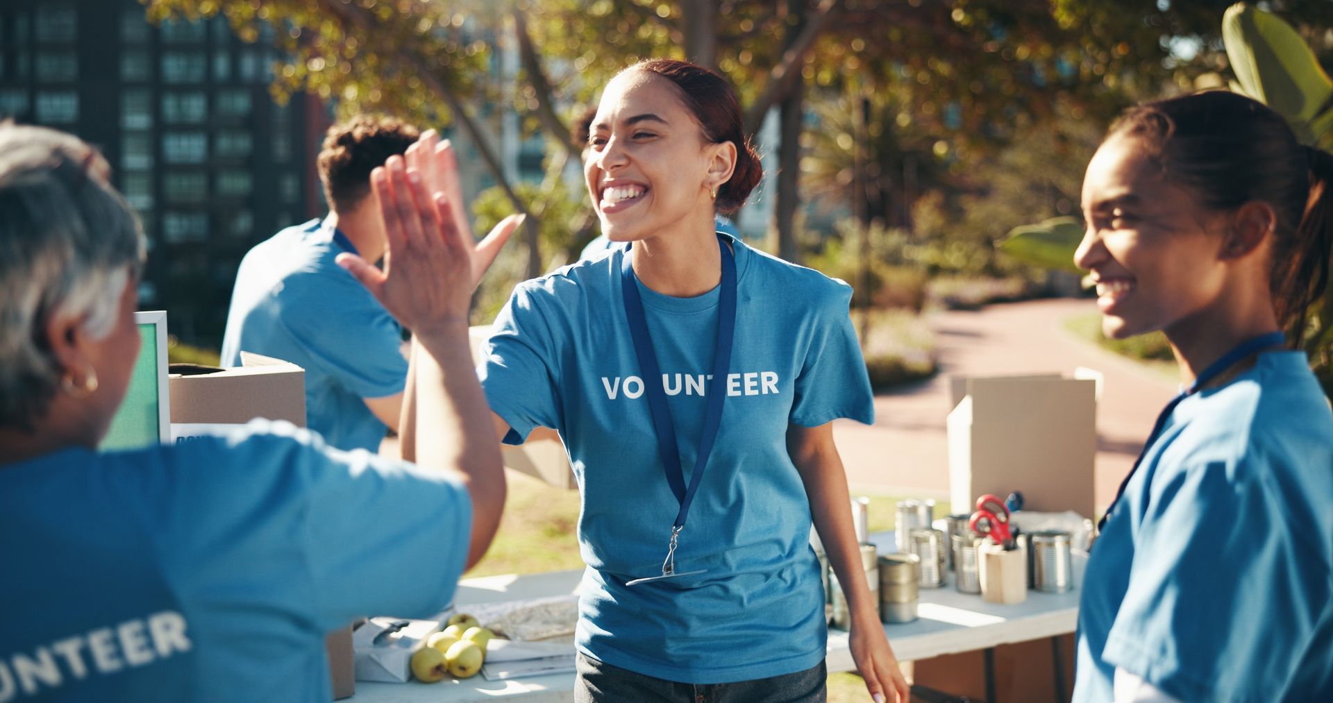 Volunteers in blue shirts high-fiving outdoors. Smiling people at a table with canned goods, sunny day.