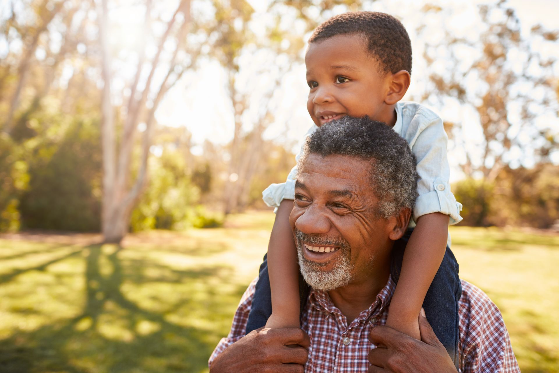Grandfather smiles as young boy sits on his shoulders in a sunny park.