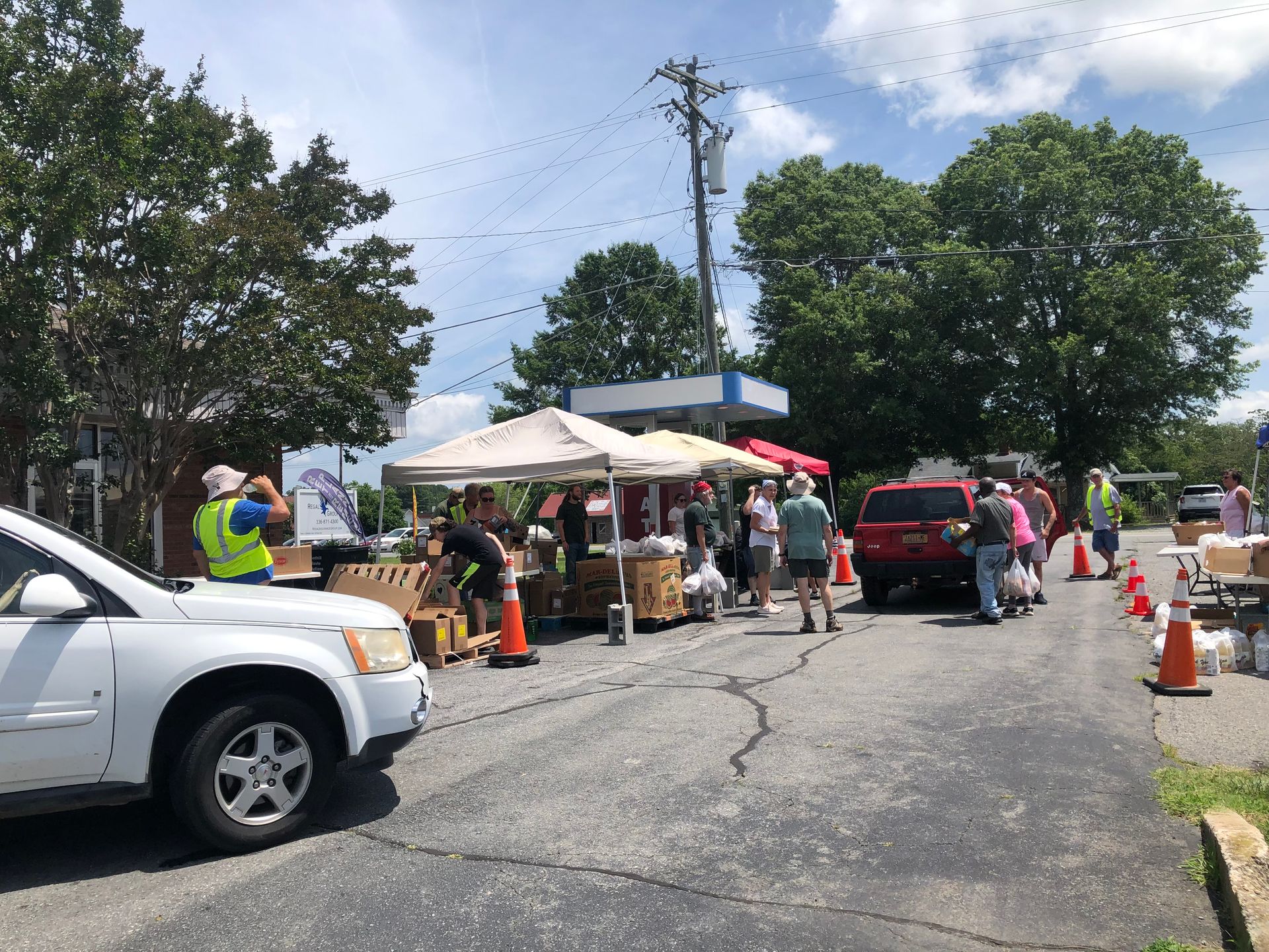 Outdoor market with tents and people browsing, cars parked, and a utility pole under a partly cloudy sky.