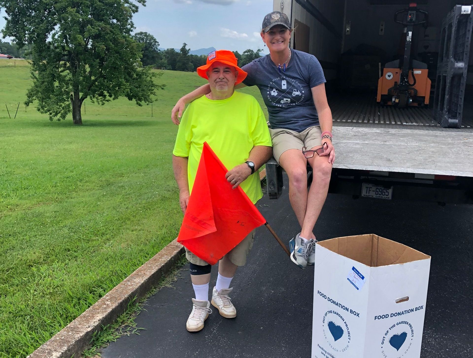 Two people pose near a truck. One holds a red flag and wears a neon shirt and orange hat. The other sits on the truck, smiling.