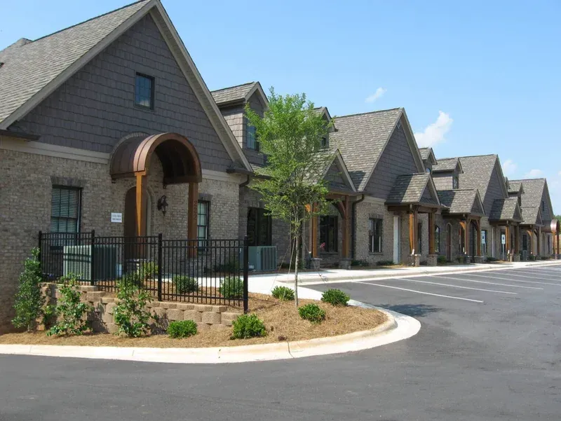 Row of brick buildings with gabled roofs and small porches on a sunny day. A black fence and shrubs are in front.