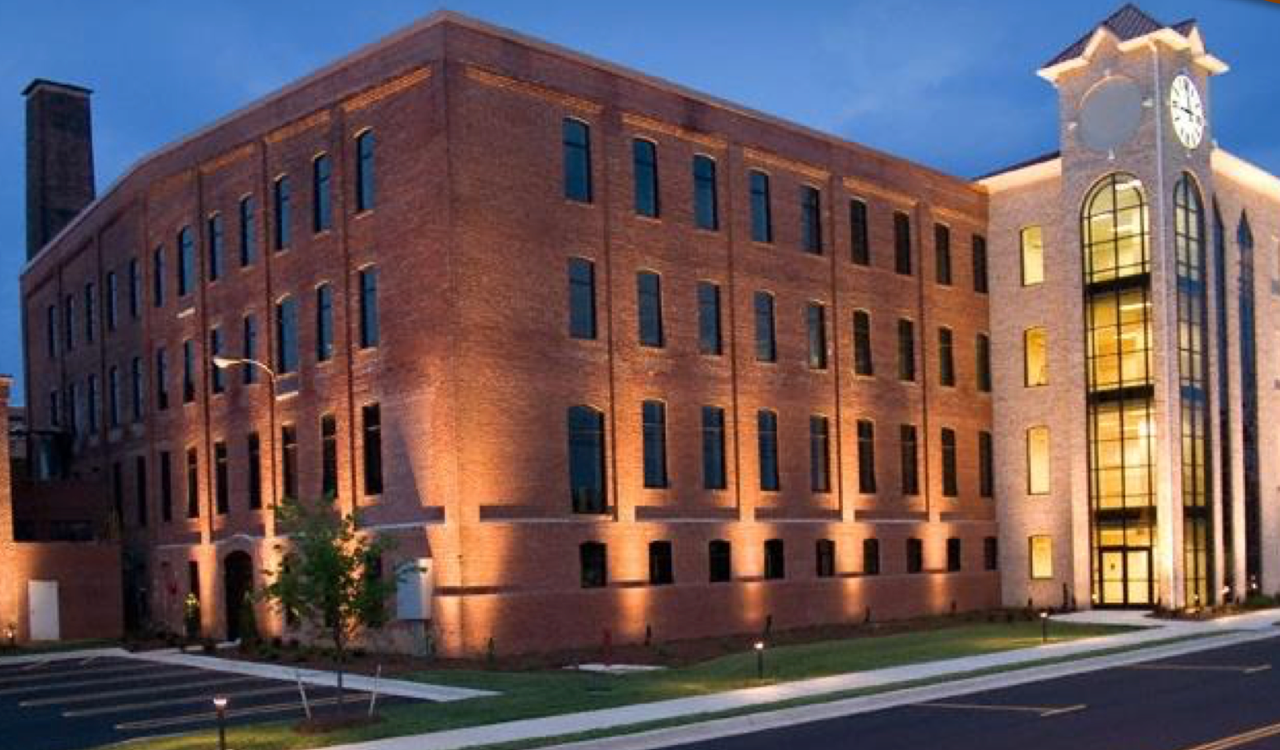 Row of brick buildings with gabled roofs and small porches on a sunny day. A black fence and shrubs are in front.