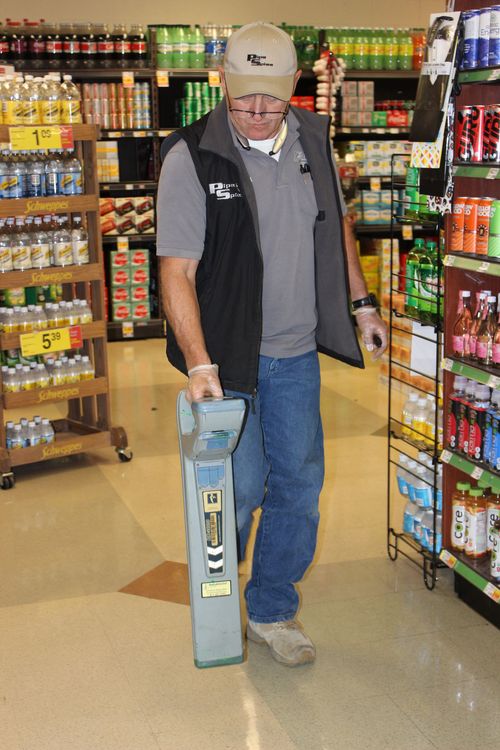 Man in store using a metal detector on the floor between shelves filled with products.