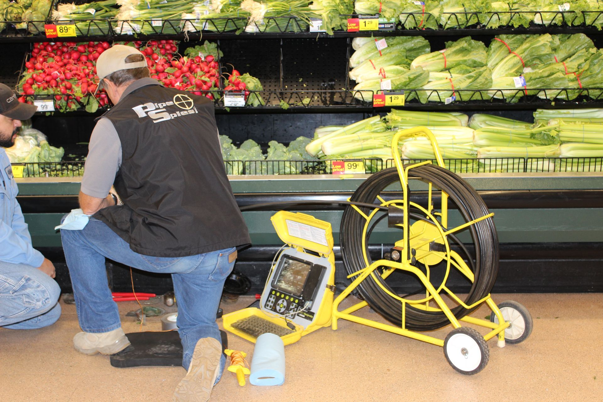 Two men with drain inspection equipment in a grocery store produce section.