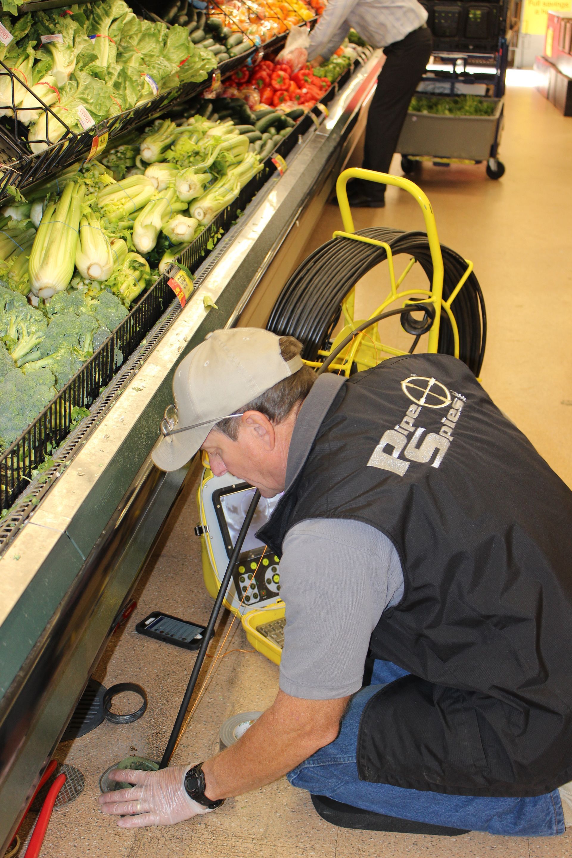 Man in a vest inspects a grocery store floor with a camera, produce in the background.