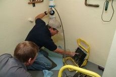 Two men in a room with plumbing, using a camera to inspect a pipe. One points at the monitor.