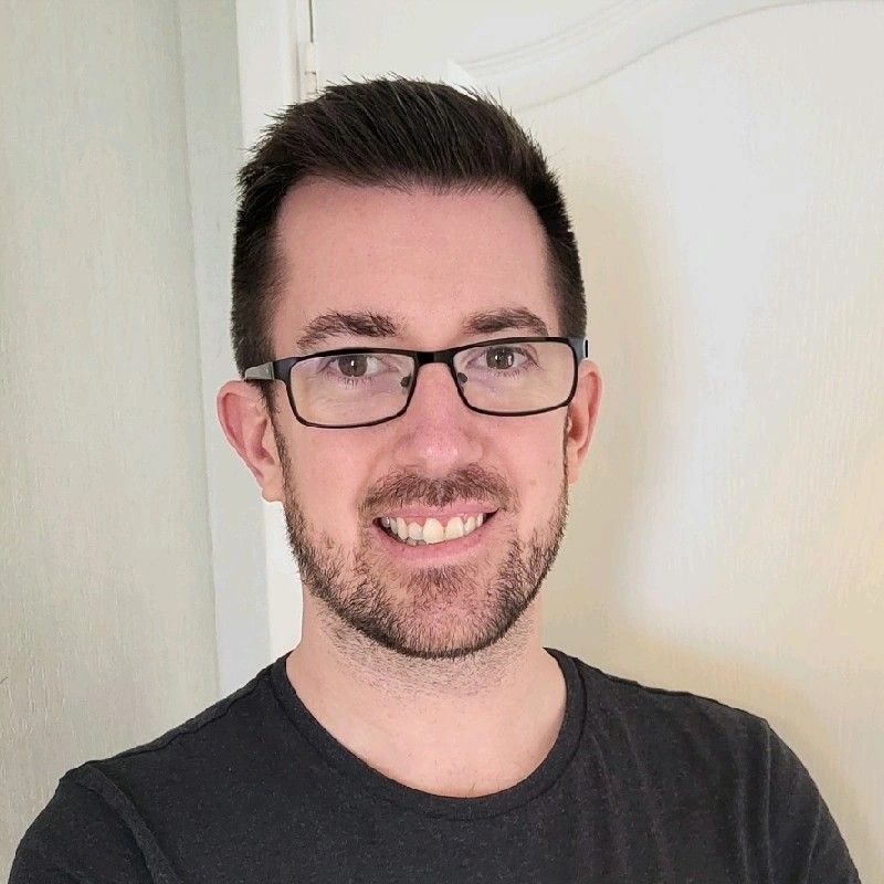 Man with glasses smiling, wearing a black shirt. Set against a white wall.