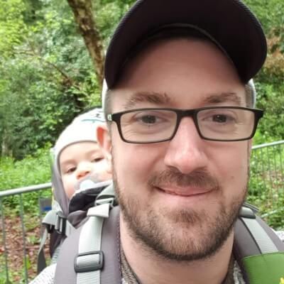 A man wearing glasses and a hat is carrying a baby in a backpack.