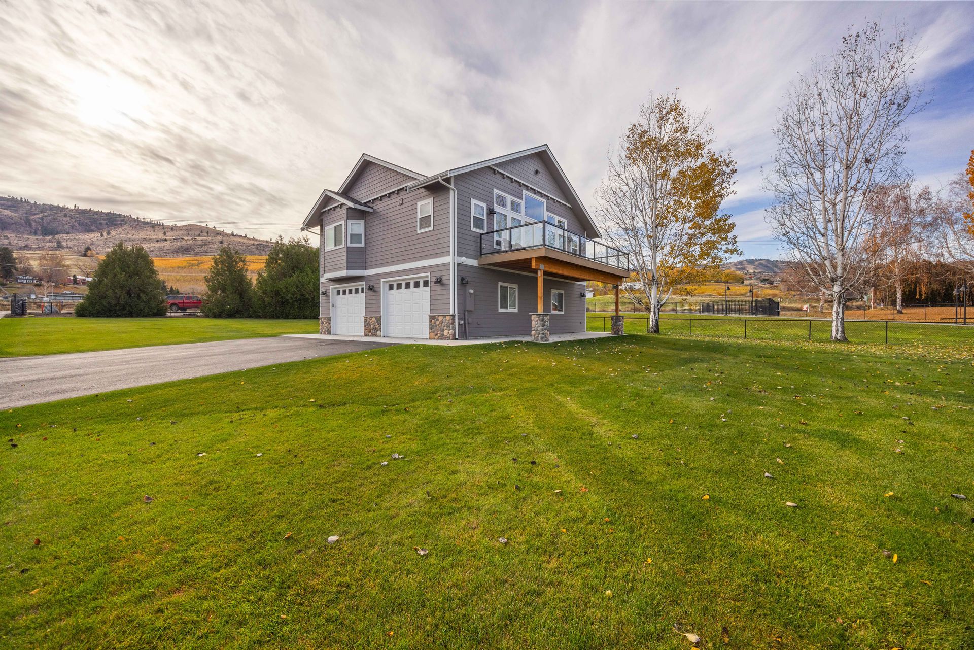 Two-story gray house with a deck and two-car garage on a grassy lawn with mountains in the background.