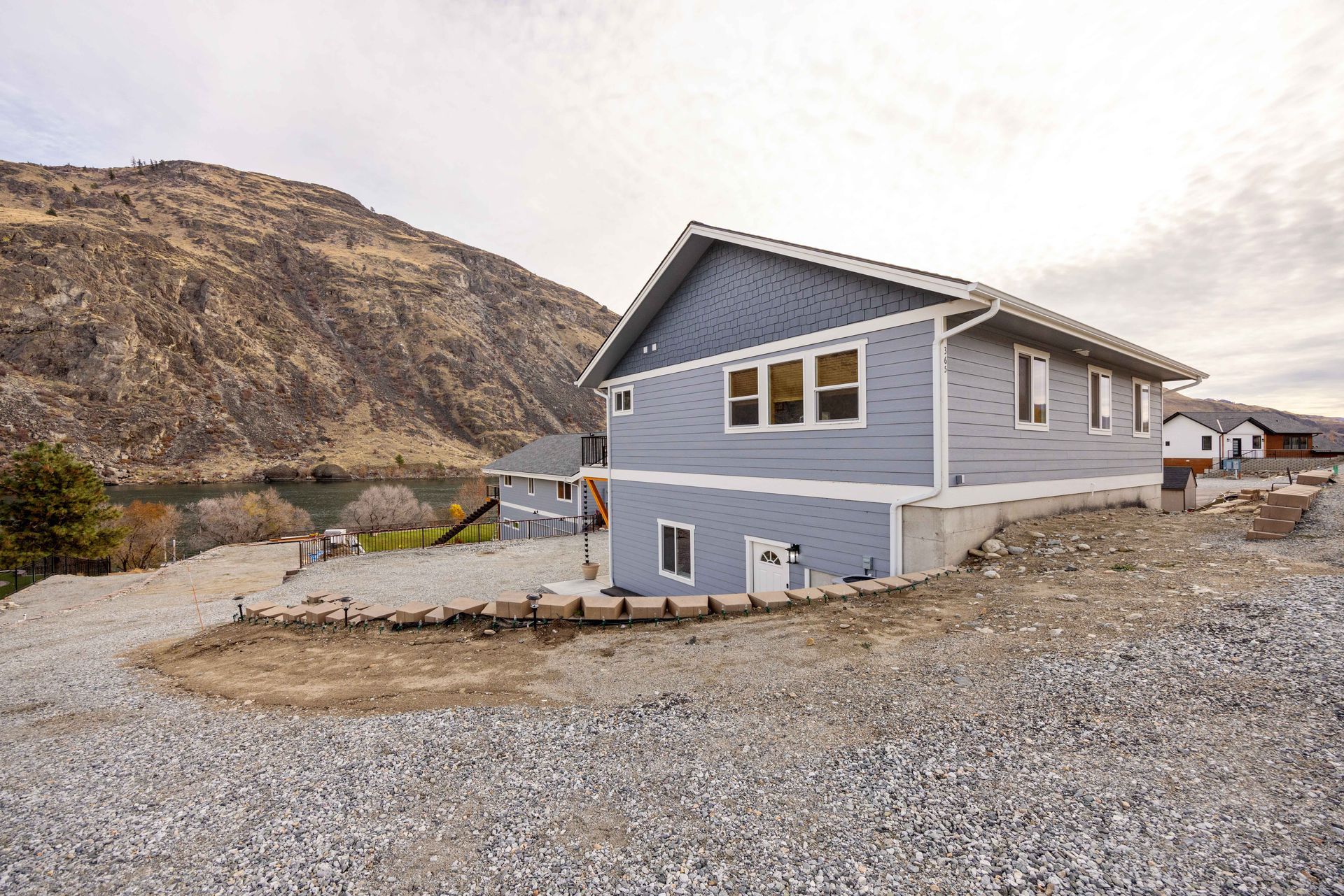 Two-story blue house on a rocky lot with a mountain backdrop under a cloudy sky.