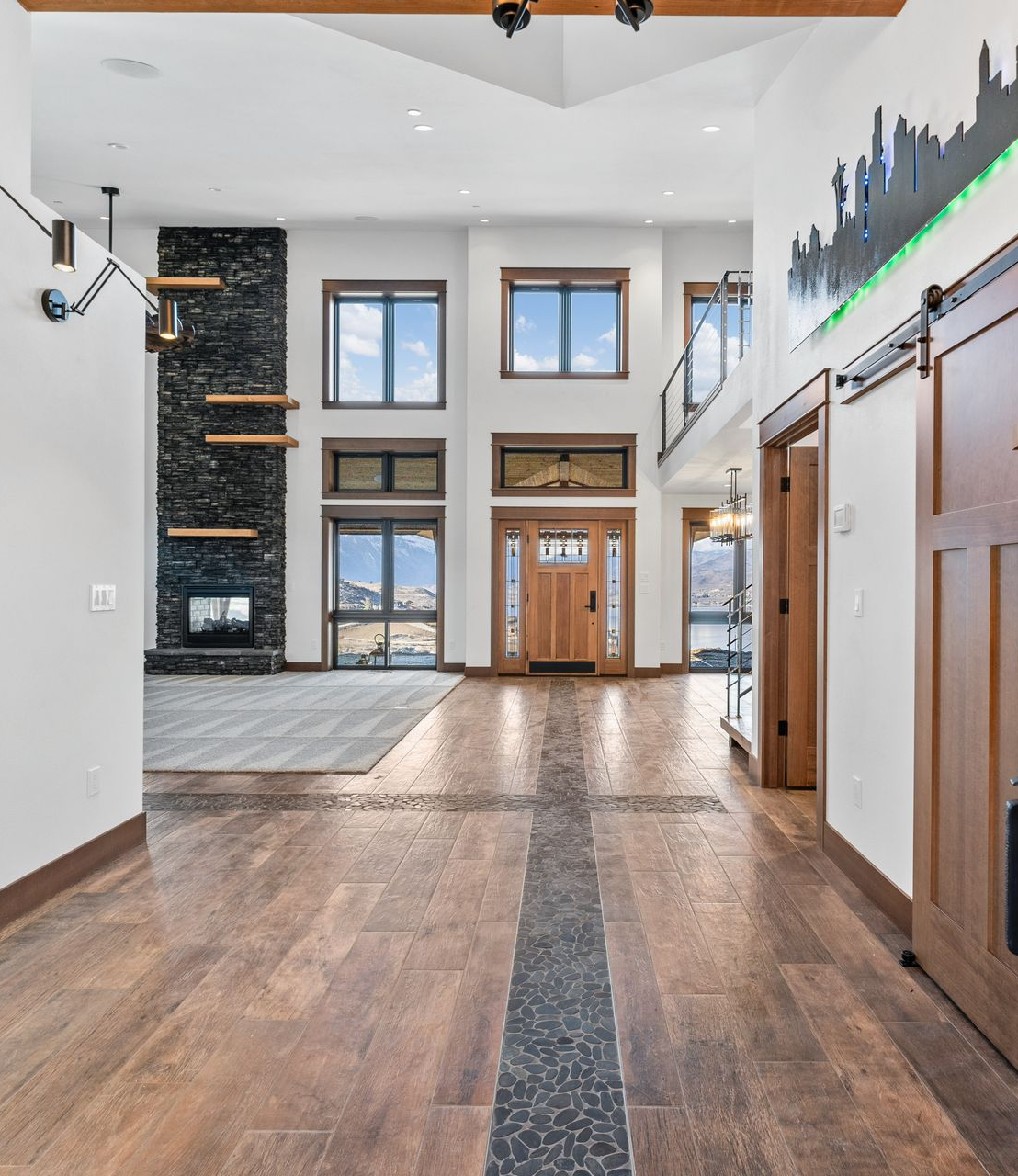 Spacious home interior: wood floors, stone fireplace, large windows, open layout, bright lighting, wood sliding door with art.