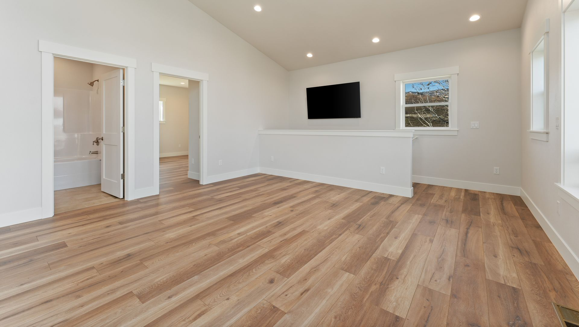 Bright bedroom with wood floor, white walls, TV, bathroom door, and window.