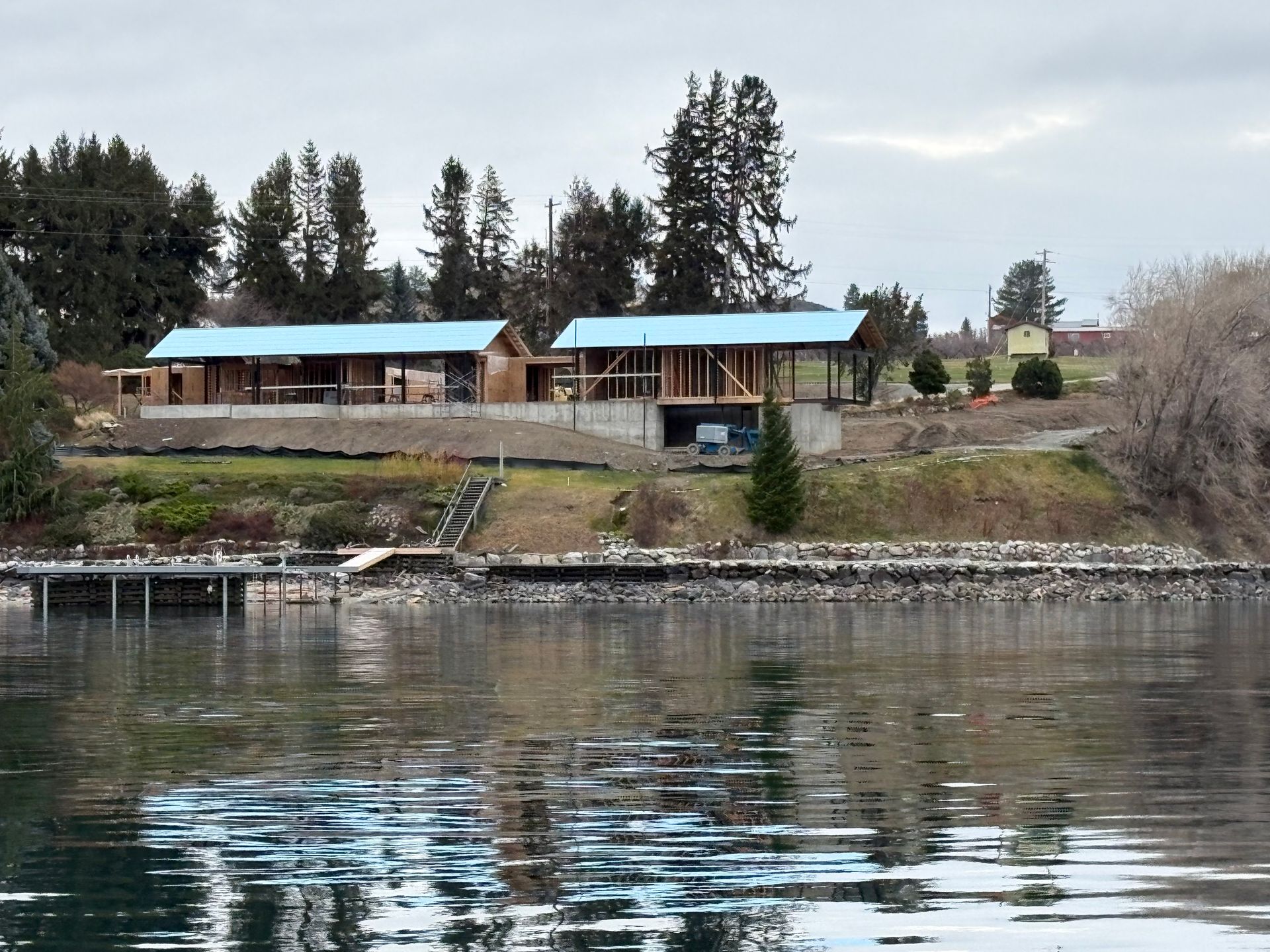 House under construction by water with blue tarp roof, overcast sky.