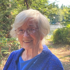 Elderly woman with white hair and glasses smiles outdoors, wearing a blue sweater.
