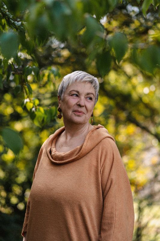 Femme aux cheveux gris courts portant un haut couleur rouille, regardant à droite, avec un arrière-plan d'arbre vert flou.