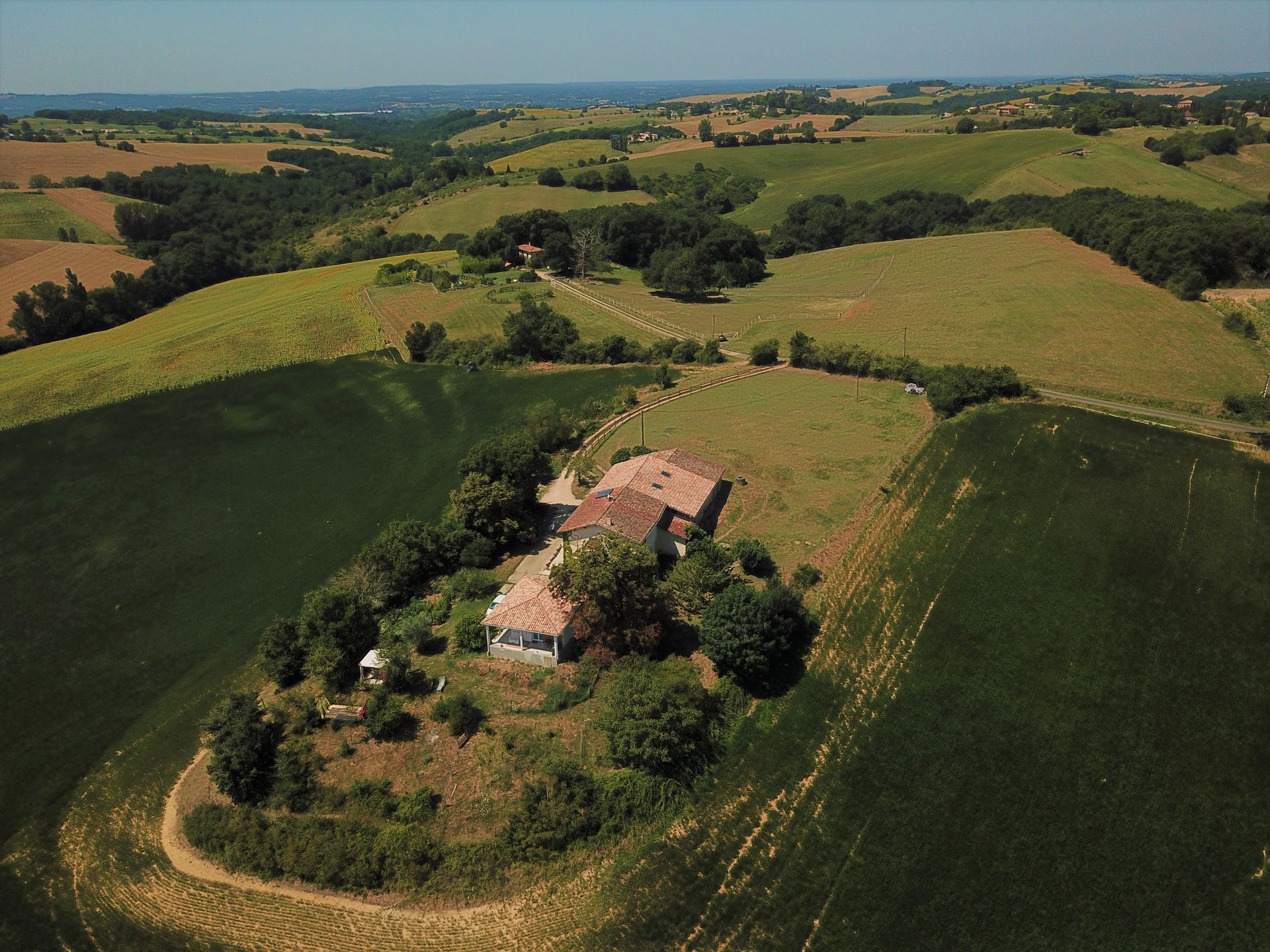 Vue aérienne d'un bâtiment rustique avec un toit de tuiles rouges entouré de collines verdoyantes et d'arbres par une journée ensoleillée.