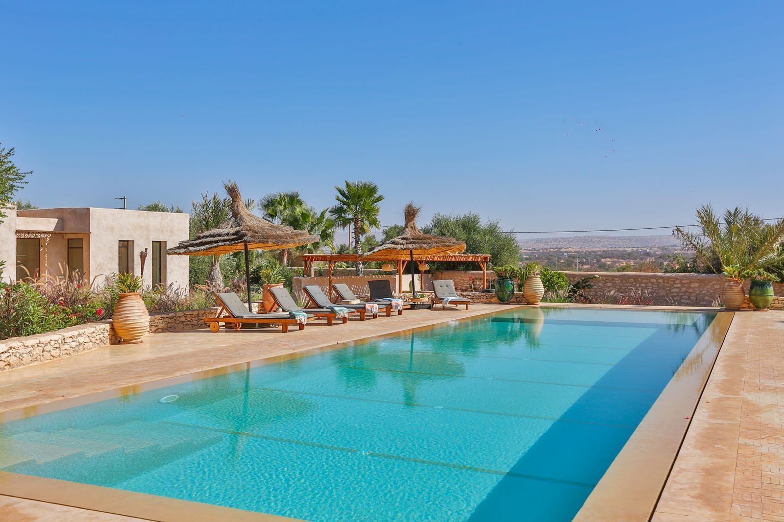 Vue sur la piscine avec chaises longues, pergolas et ciel bleu clair.