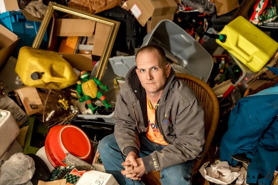 A man sits in a chair surrounded by cluttered piles of boxes, household items, a yellow fuel jug, and a toy action figure.