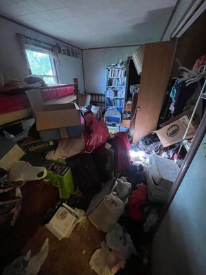 A cluttered bedroom with a bed, a tall shelf filled with items, and numerous boxes and bags scattered across the floor.