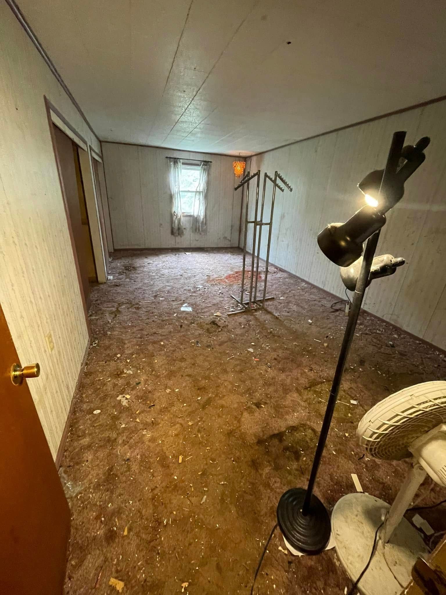 A vacant room with white walls, a single window, a metal garment rack, a floor lamp, and scattered debris on the carpet.