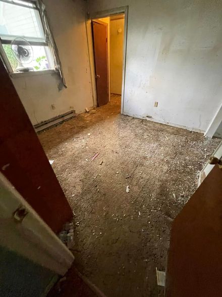 A small, empty room with debris scattered across a dark floor, white walls, and a window with a fan.