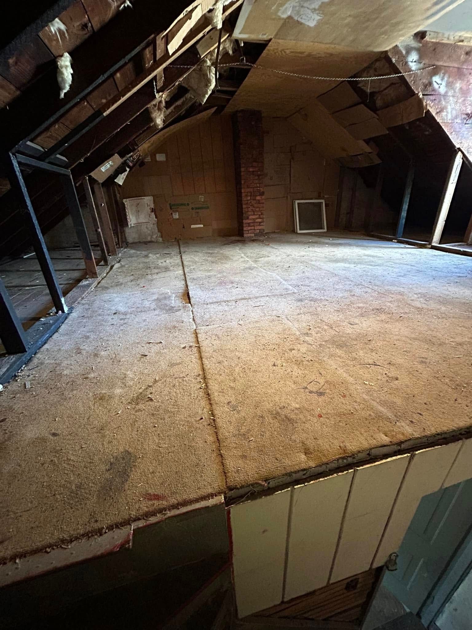 An unfinished attic with exposed wooden rafters, a brick chimney, and plywood subflooring spanning the floor.