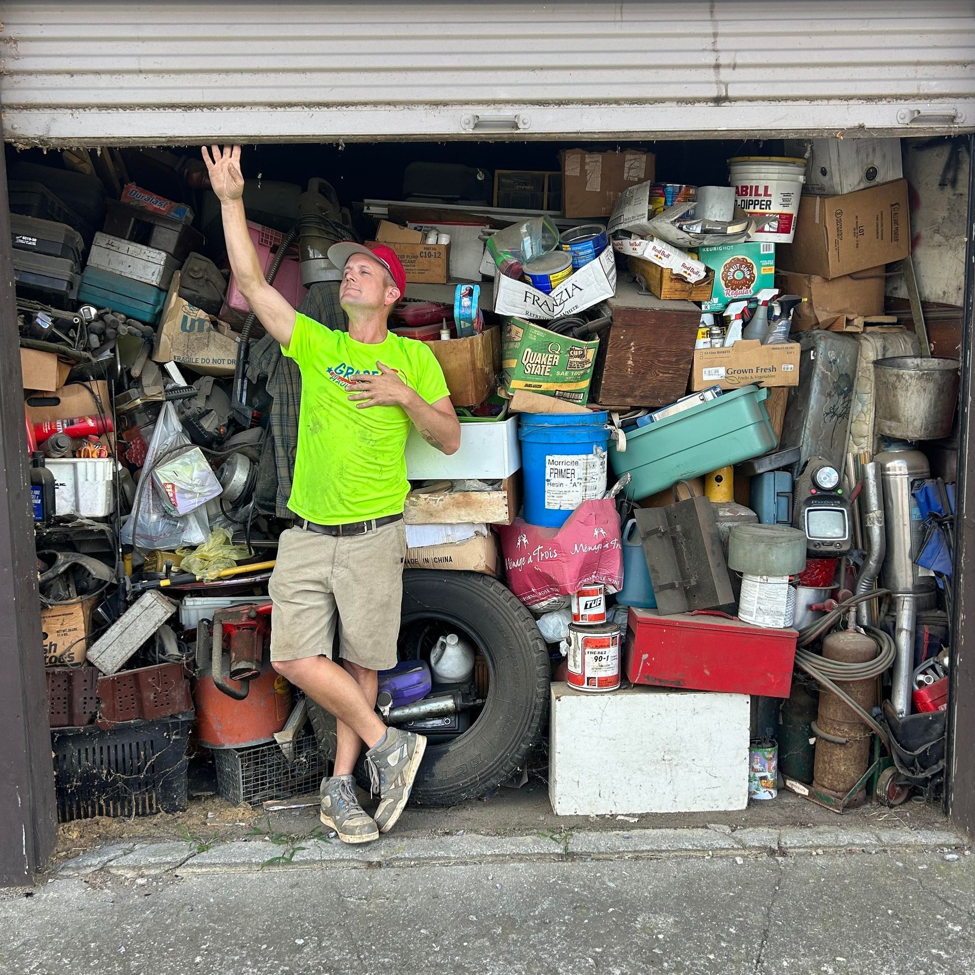 A person in a neon green shirt and shorts leans against a large tire inside a storage unit packed with miscellaneous items.