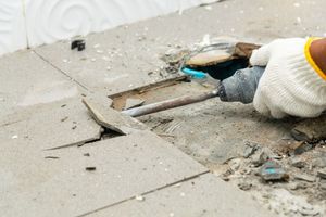A gloved hand uses a power chisel to remove gray floor tiles in a home renovation setting.