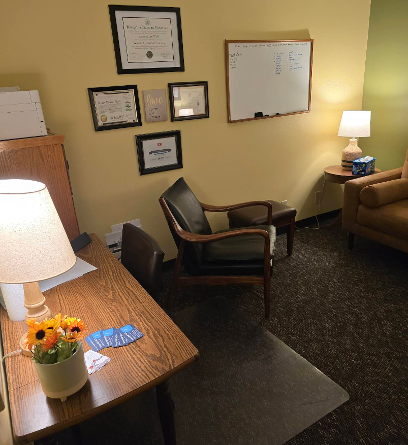 A therapist's office with a desk, chairs, artwork on the wall, and a lamp. The room is painted yellow and green.