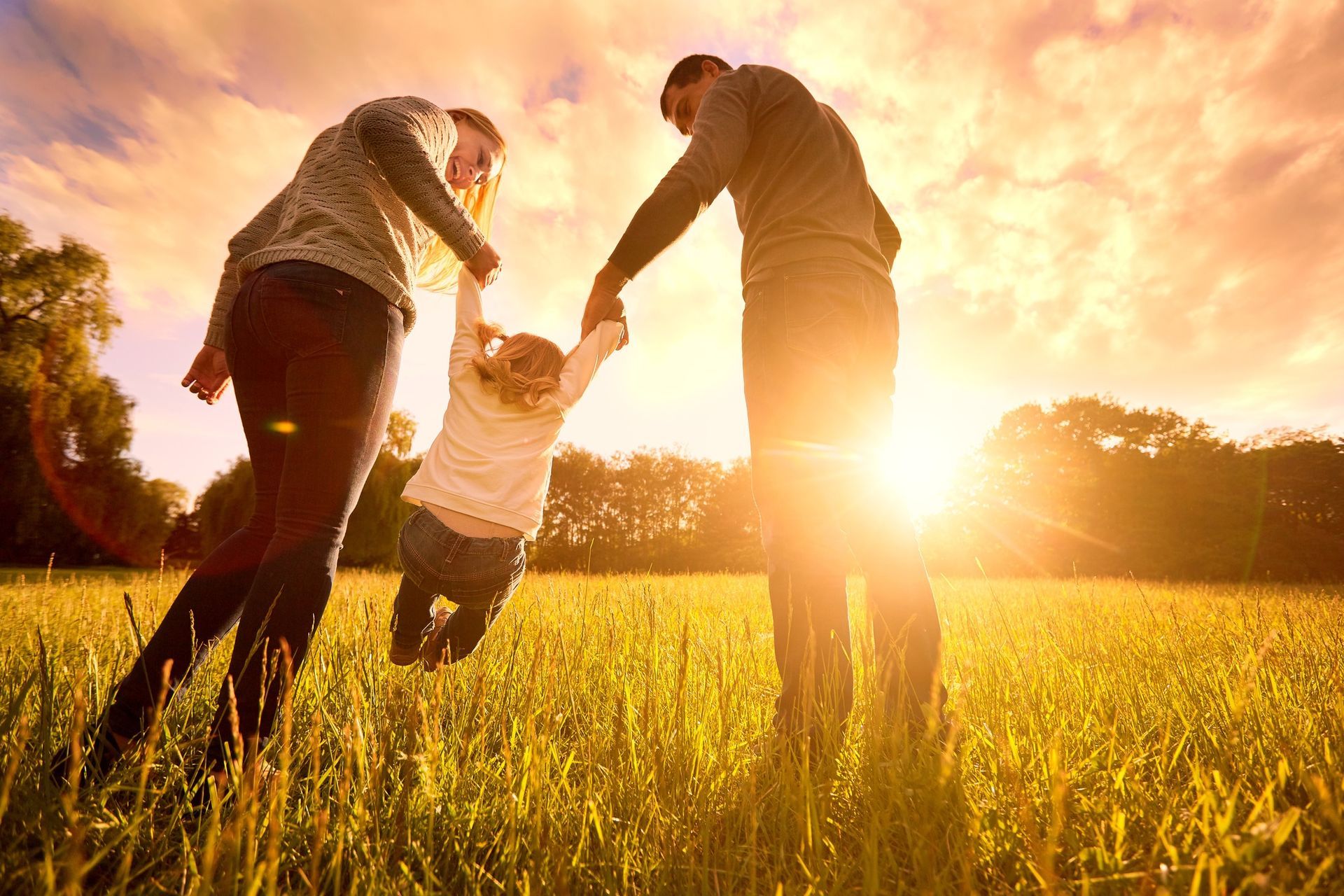 Parents swinging a child in a sunlit field; backlit figures and bright, golden light.