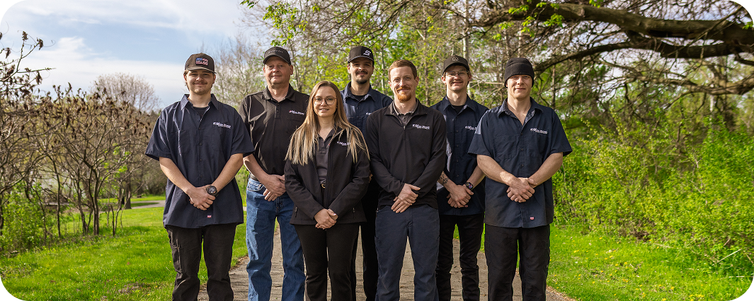 Seven people in dark clothing standing together in a green orchard under trees | Bob's Main Street Auto