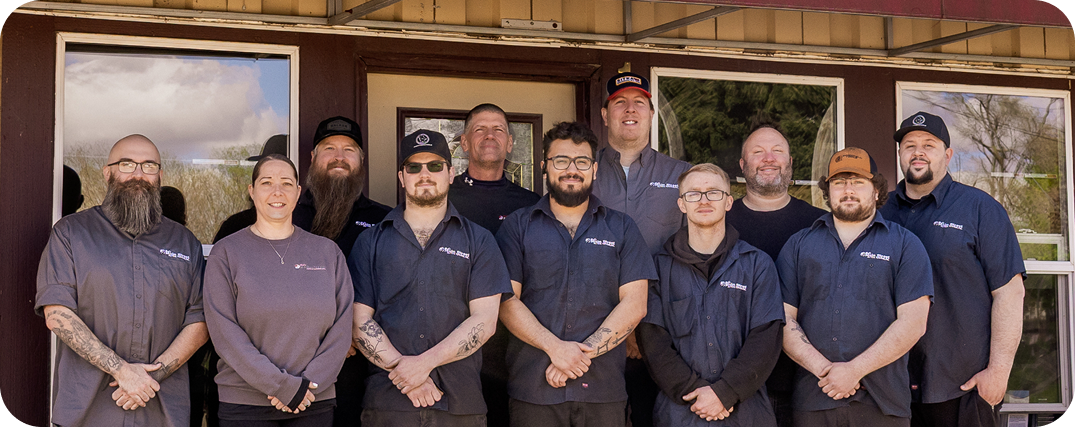 Group of eight people standing outside in front of a light-colored wall, smiling at the camera. | Bob's Main Street Auto & Towing