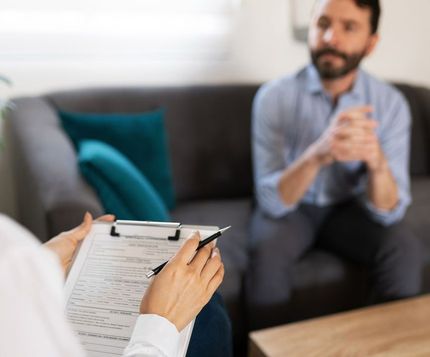 A woman is holding a clipboard and talking to a man sitting on a couch.