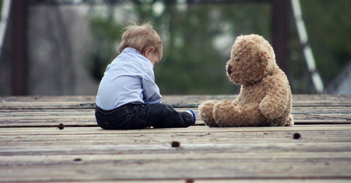 A little boy is sitting next to a teddy bear on a wooden deck.