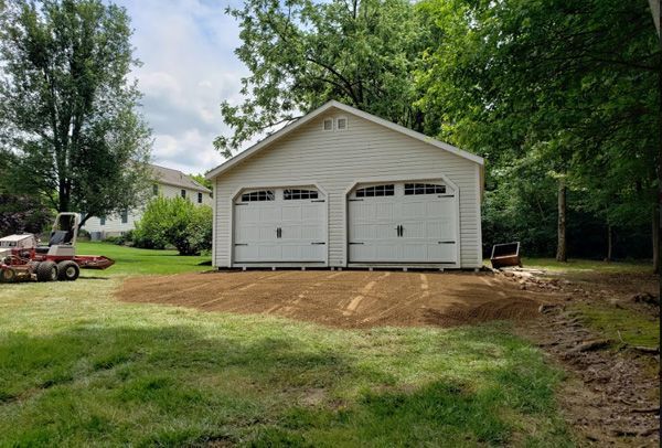 A white garage with two garage doors is sitting in the middle of a lush green field.