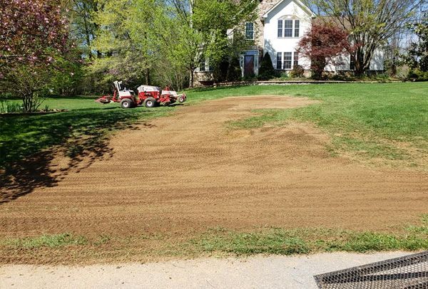 A red tractor is driving down a dirt road in front of a house.