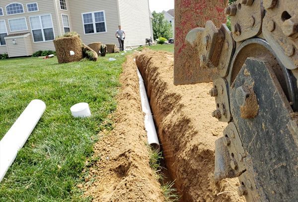 A man is standing in the grass looking at a trench being dug in front of a house.