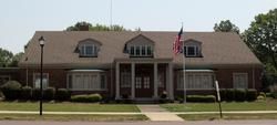 A symmetrical brick building with a gabled roof, three dormer windows, a columned porch entrance, and an American flag.