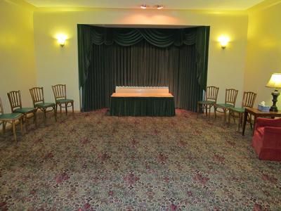 A room with patterned carpeting, a central stage with dark green curtains, and chairs arranged for a small gathering.