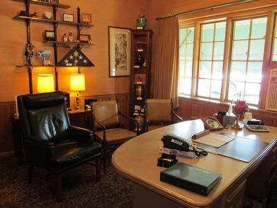 An office interior with a curved desk, a leather armchair, two guest chairs, a display case, and wall-mounted shelving.