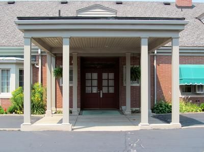 A red brick building entrance with a tan portico supported by four pillars, leading to a set of double glass doors.