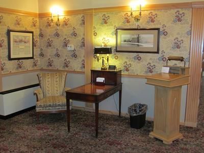 A cozy corner featuring patterned wallpaper, a velvet armchair, a small desk with a lamp, and a wooden podium.