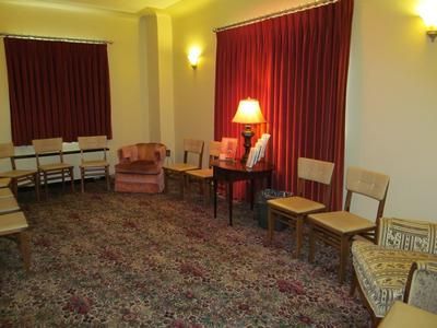 A waiting room with patterned carpet, maroon curtains, several wooden chairs, a side table with a lamp, and armchairs.