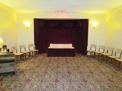 A room with a maroon curtain backdrop behind a long table, set with chairs arranged on both sides on patterned carpet.