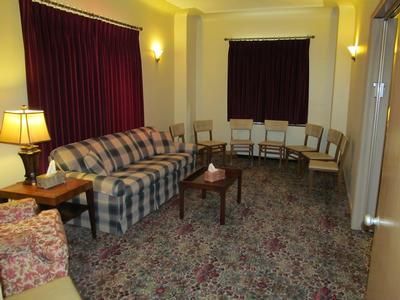 A waiting room with a plaid sofa, wooden coffee table, and several wooden chairs on a floral carpet under maroon curtains.
