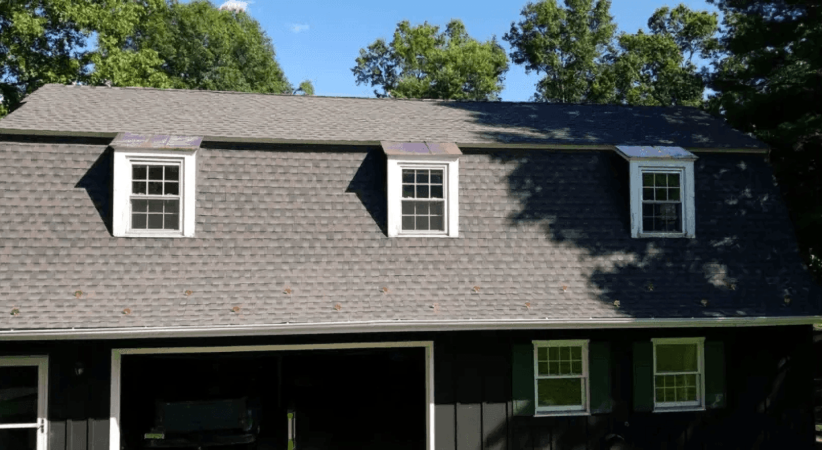 Two-story gray house with dormer windows and a dark garage under a tree-lined blue sky