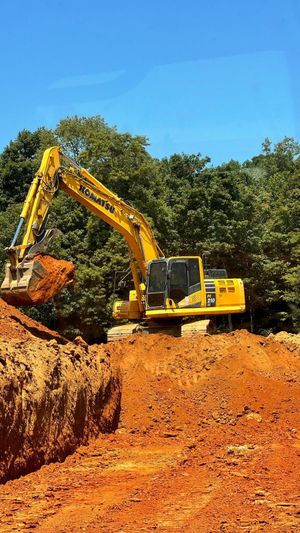 A yellow excavator digs into a large pile of reddish-brown earth against a backdrop of trees and a clear blue sky.