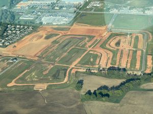 An aerial view of a construction site with freshly graded roads and dirt lots surrounded by fields and a nearby neighborhood.