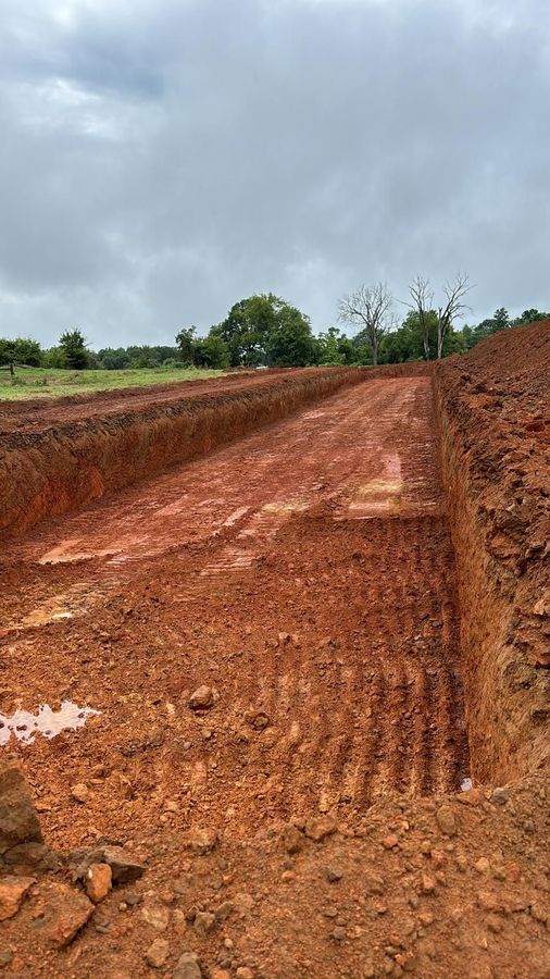 A wide, rectangular trench excavated in red clay soil under a cloudy sky.